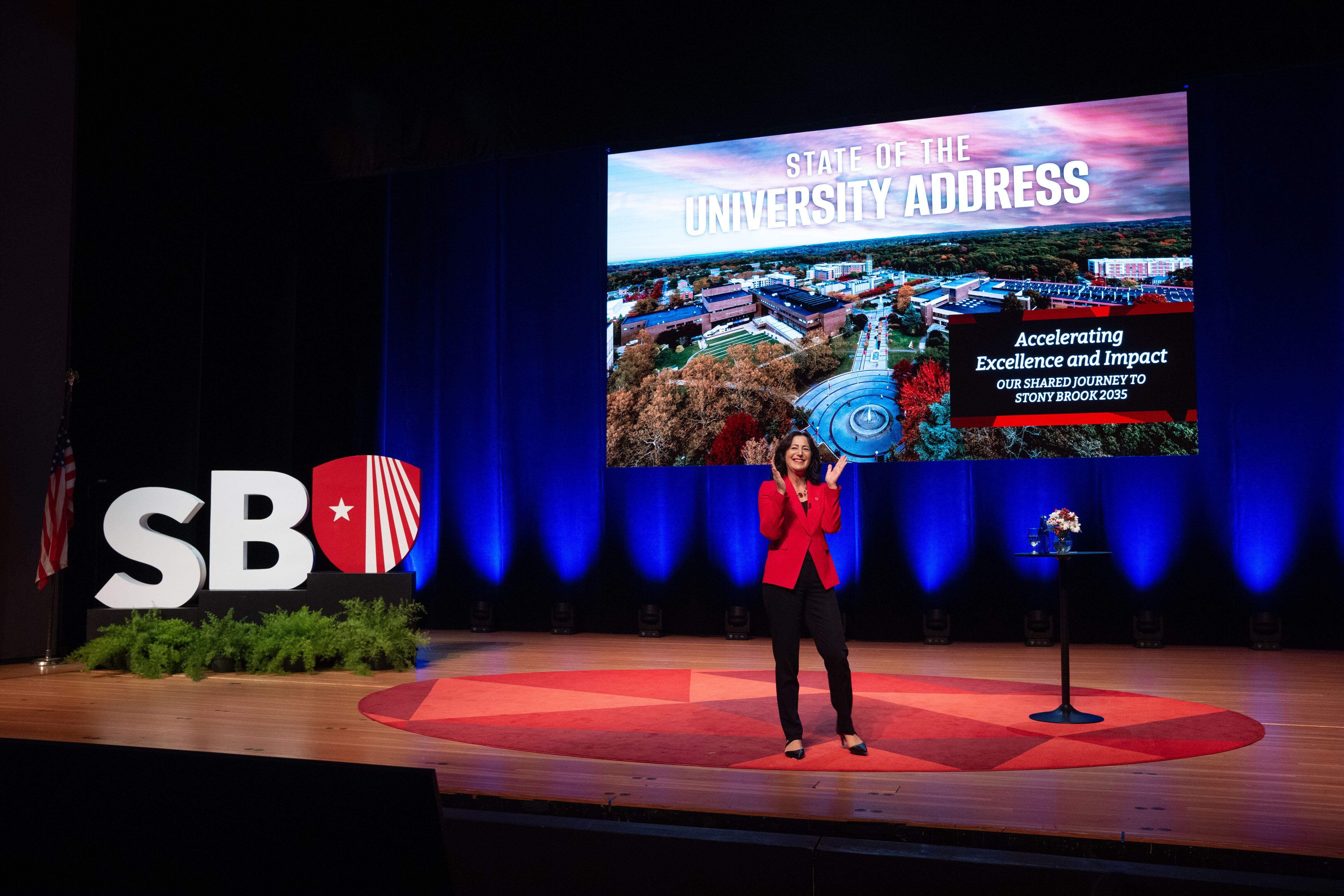 President Goldsmith in a red blazer stands on a stage in front of a screen reading "State of the University Address" next to a large "SBU" logo.