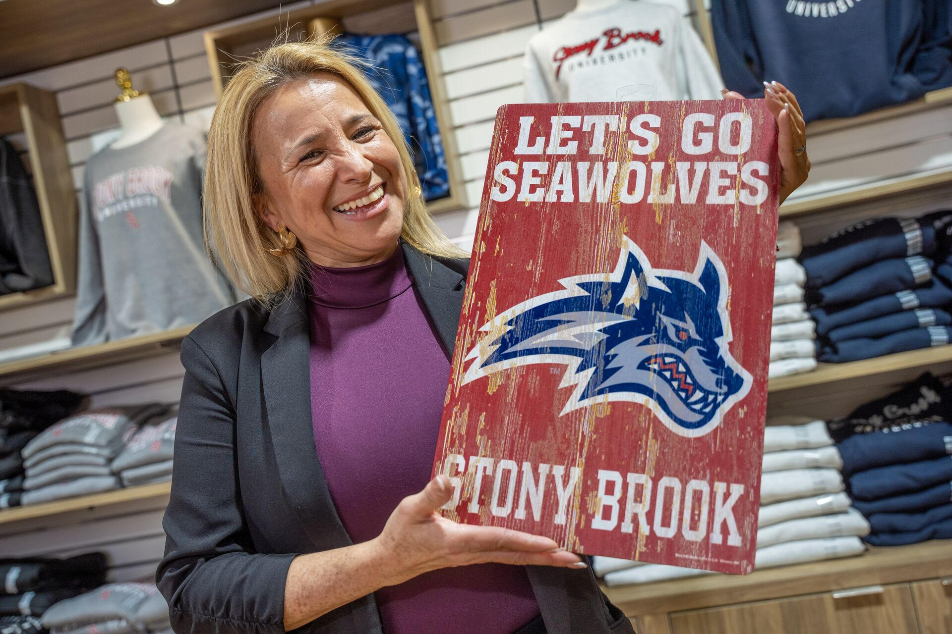 A smiling woman holds up a wooden Stony Brook sign reading "Let's Go Seawolves."