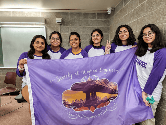 A group of six women in purple and white shirts pose in a classroom, holding a flag that reads “Society of Women Engineers.”
