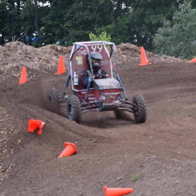 A student in a red baja style racecare drives around the curve of a dirt track lined with orange cones. 