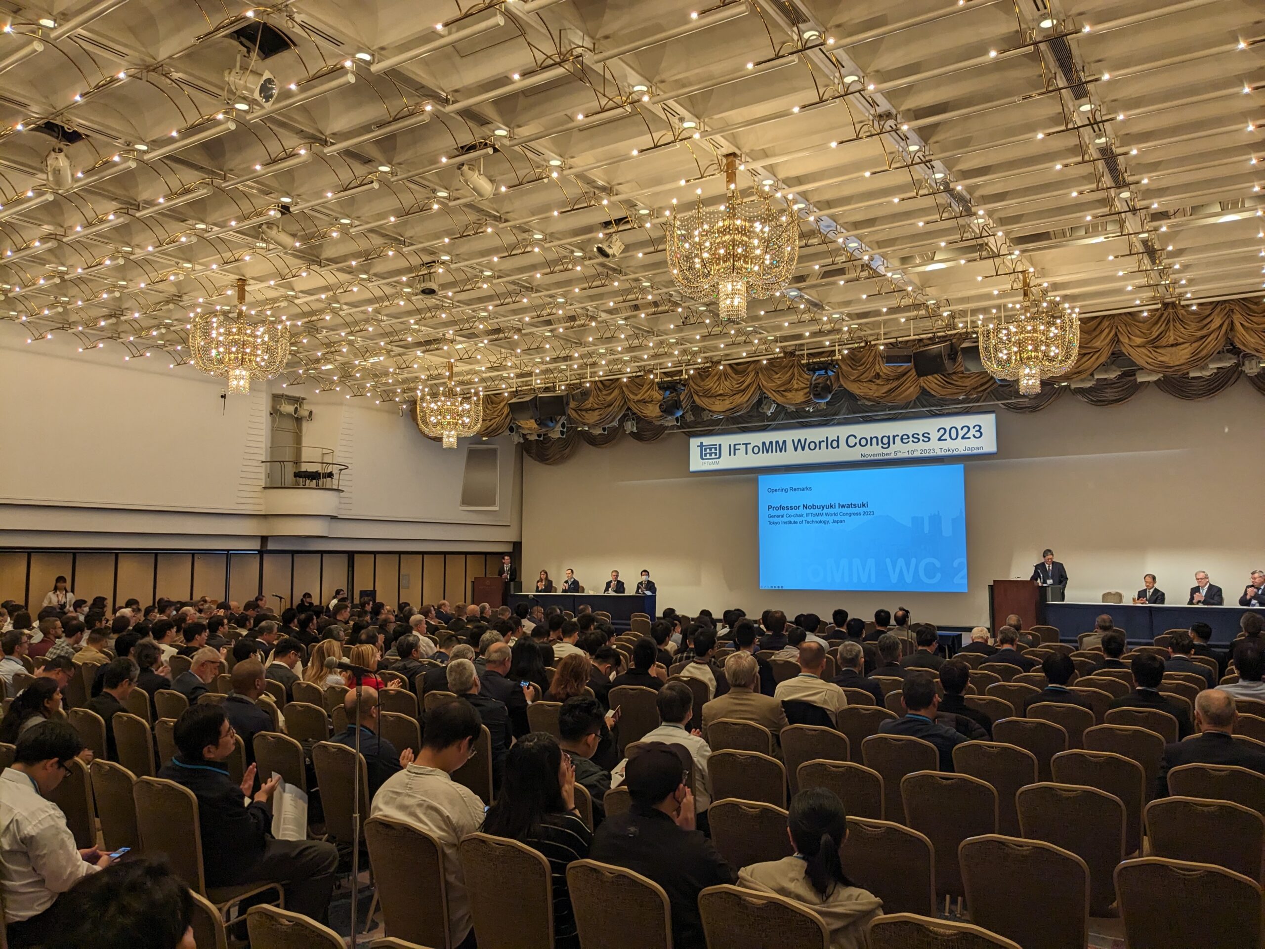 A ballroom of conference attendees with a panel of speakers seated on either side of a screen