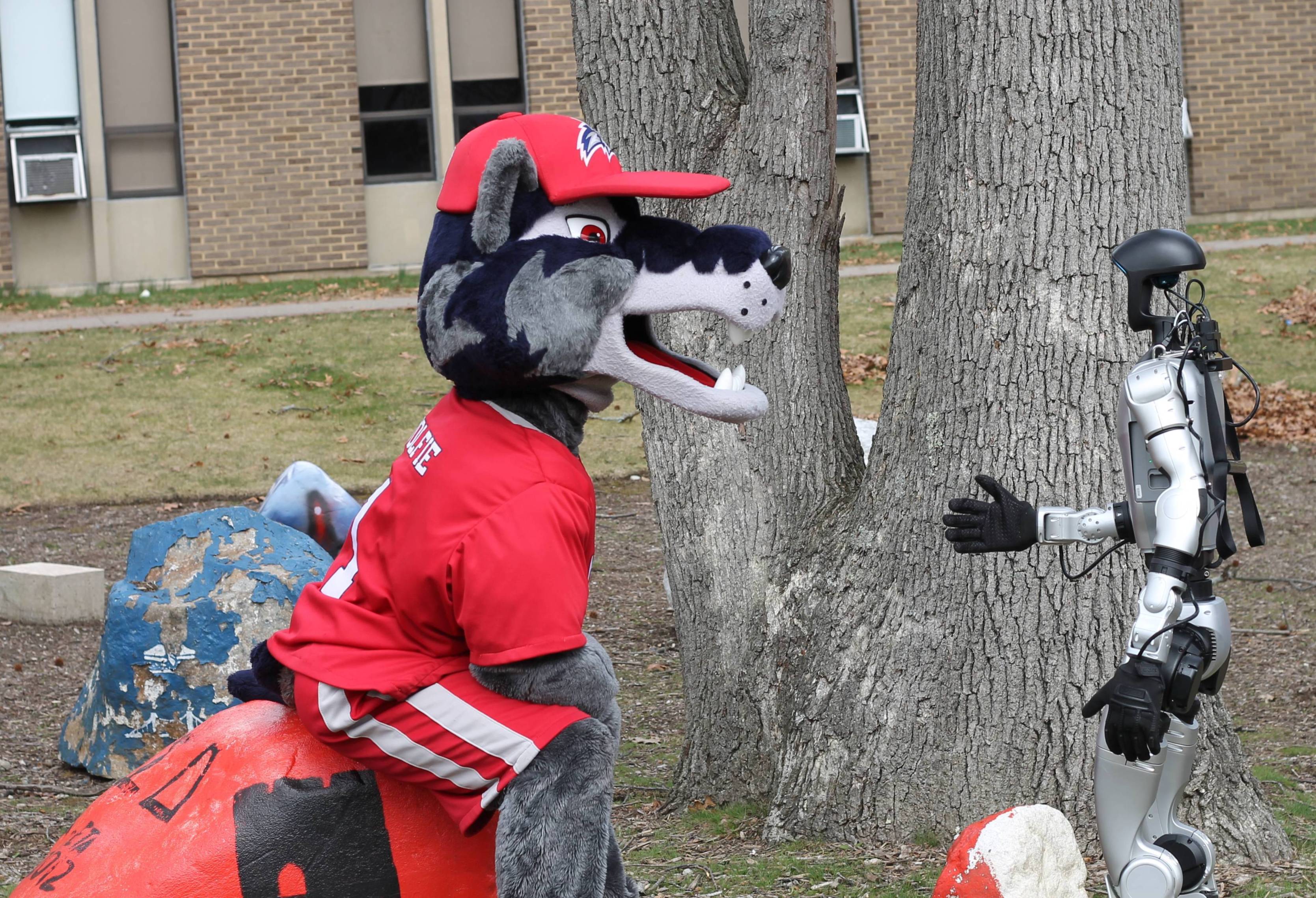 Stony Brook University mascot Wolfie the Seawolf interacting with the Department of Mechanical Engineering's robot in front of the engineering building. 