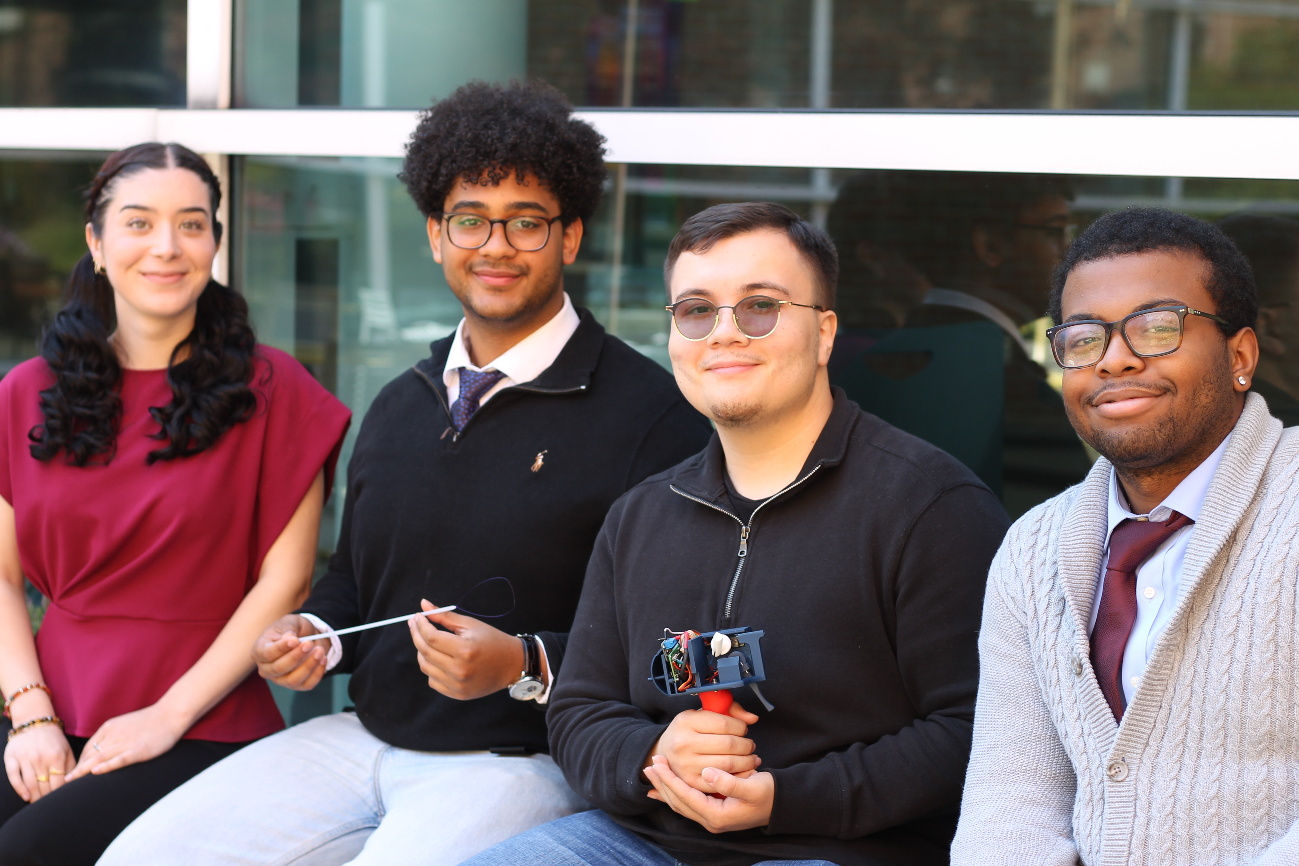 Four people seated in front of a glass building; one holds a small wired electronic device.