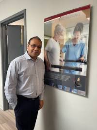 Professor Purwar poses next to a plaque featuring Mobility Assist on the Wall of Innovation