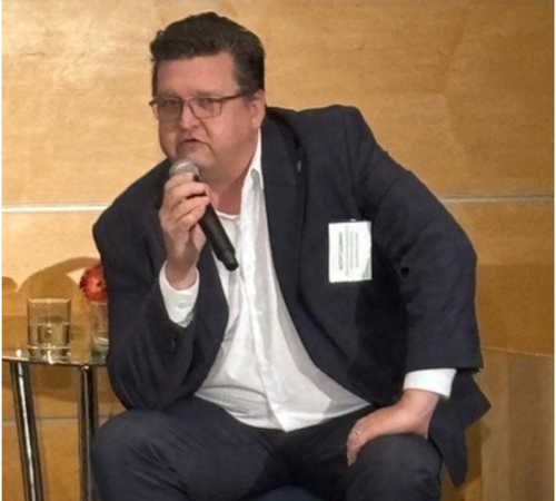 P. Scott Carney in a dark blazer speaks into a microphone at a panel discussion during the Beyond AI Quantum event.