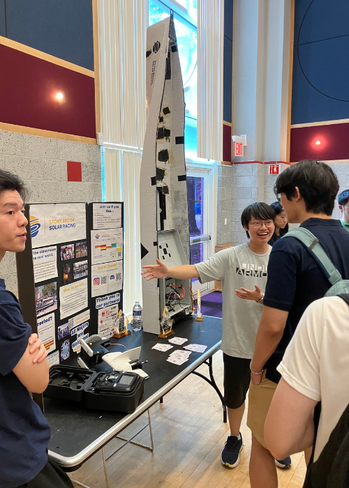 Students discuss the Stony Brook Solar Racing Team beside a tall solar race-boat prototype at a campus involvement fair.