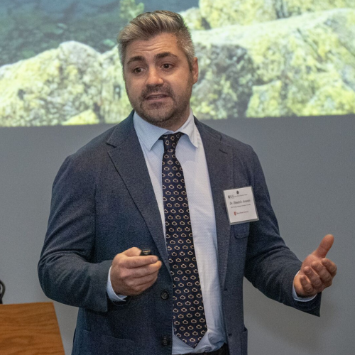 Professor Dimitris Assanis wears a navy blazer, light blue shirt, and patterned tie while speaking in front of a projection screen at a Young Academic Inventor Symposium.