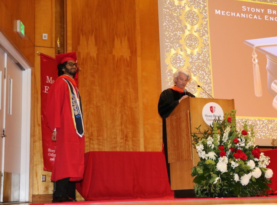 JAson Hamechand in cap and gown walks the stage at graduation with Professor Stewart at the podium