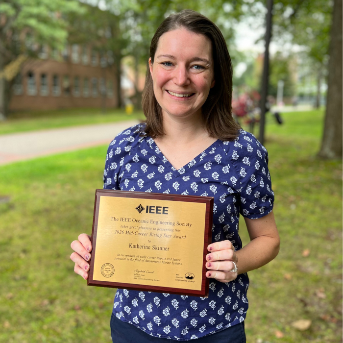 Smiling woman in a navy floral blouse holds an IEEE Oceanic Engineering Society award plaque outdoors.