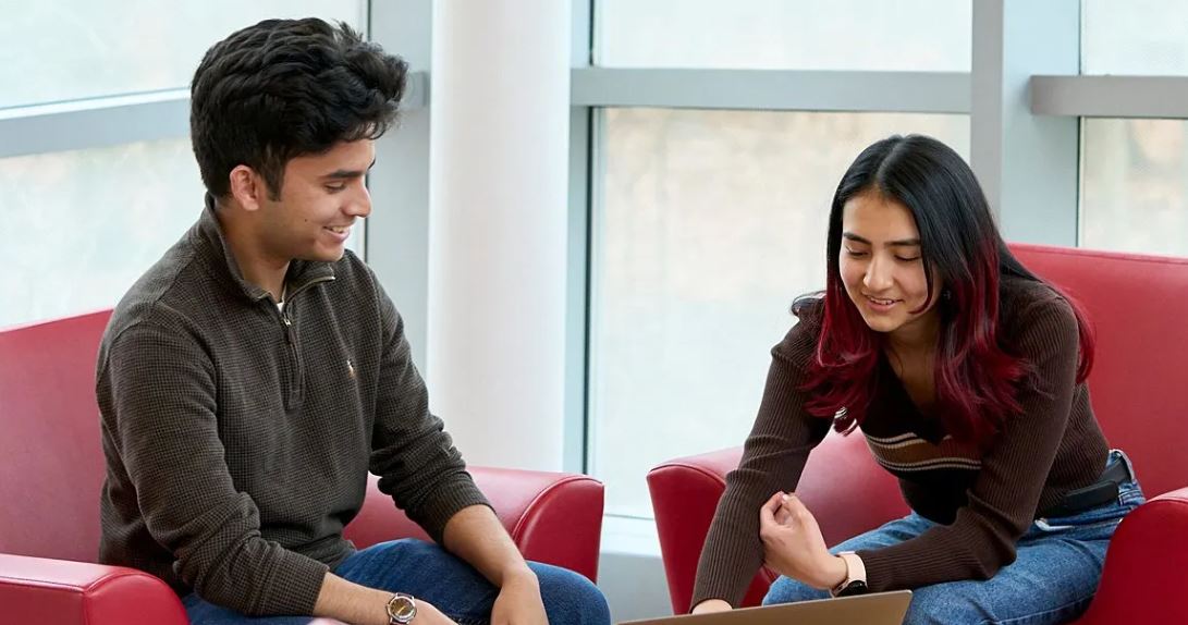 two students seated in red chairs look at a laptop