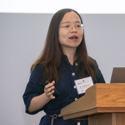Professor Shanshan Yao wearing a dark blue shirt speaks at a wooden podium