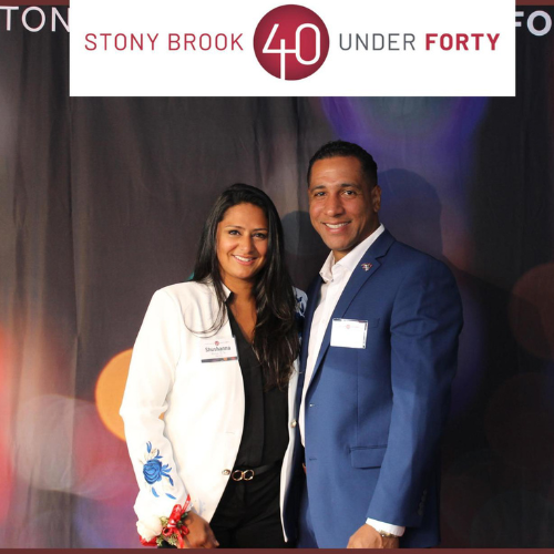 Stony Brook 2026 40 Under Forty  Honoree, Shushanna   Cherian-Brutus and her husband, Jean Clifford Brutus pose together beneath a banner that says Stony Brook “40 Under Forty”.