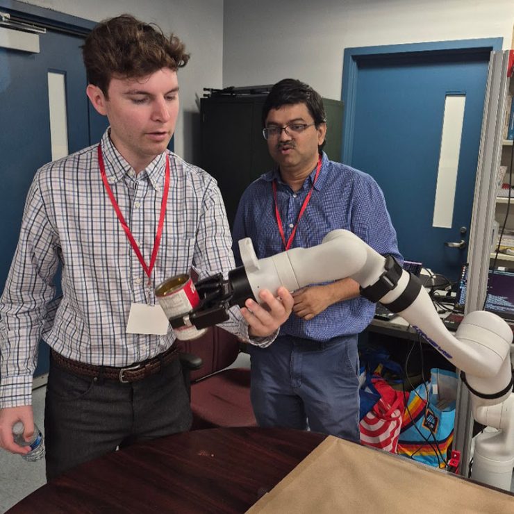 Two men demonstrate a white robotic arm holding a small can in a laboratory.