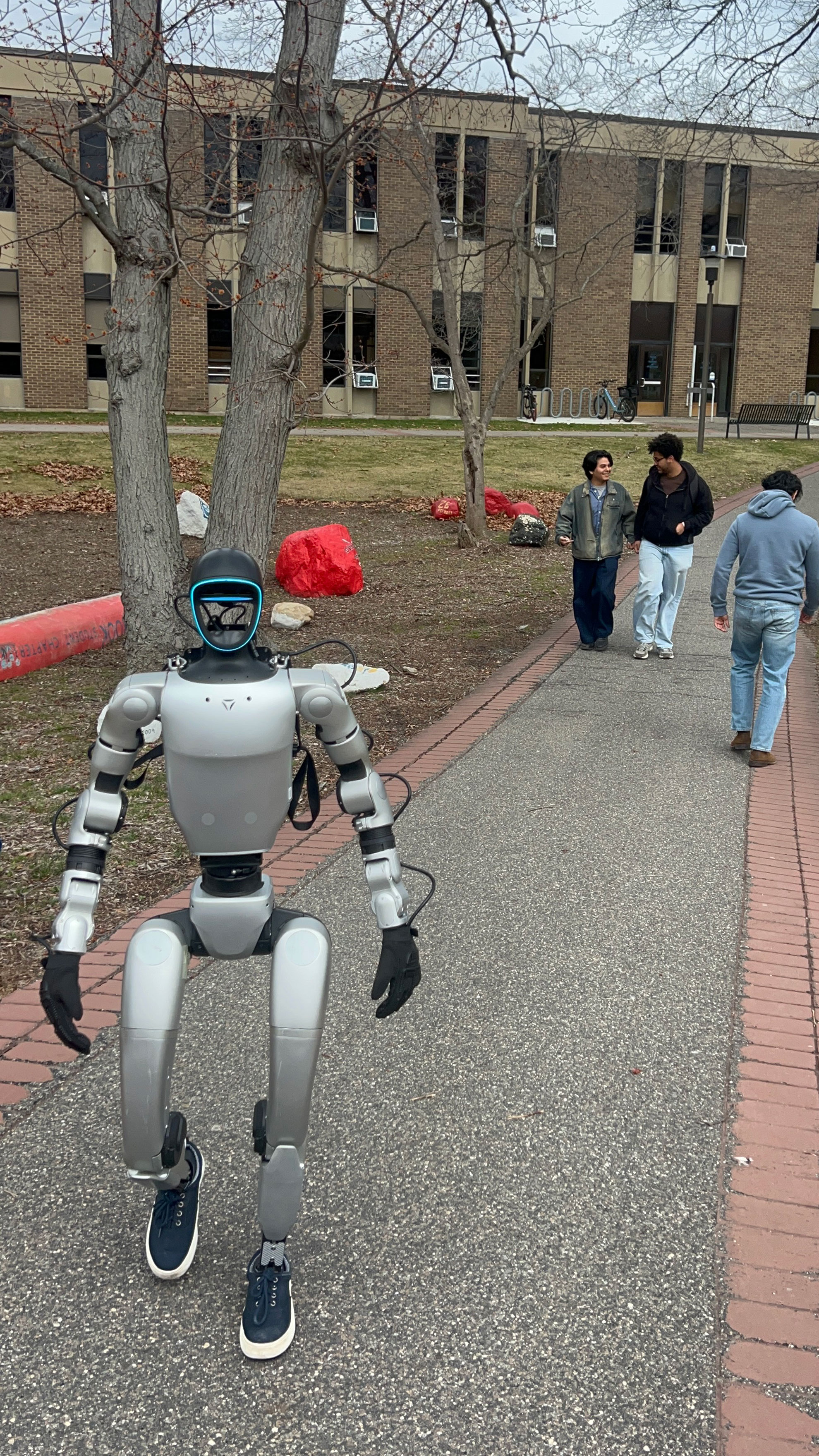 A humanoid robot stands on a walkway in campus scenery, while three people walk together in the background.