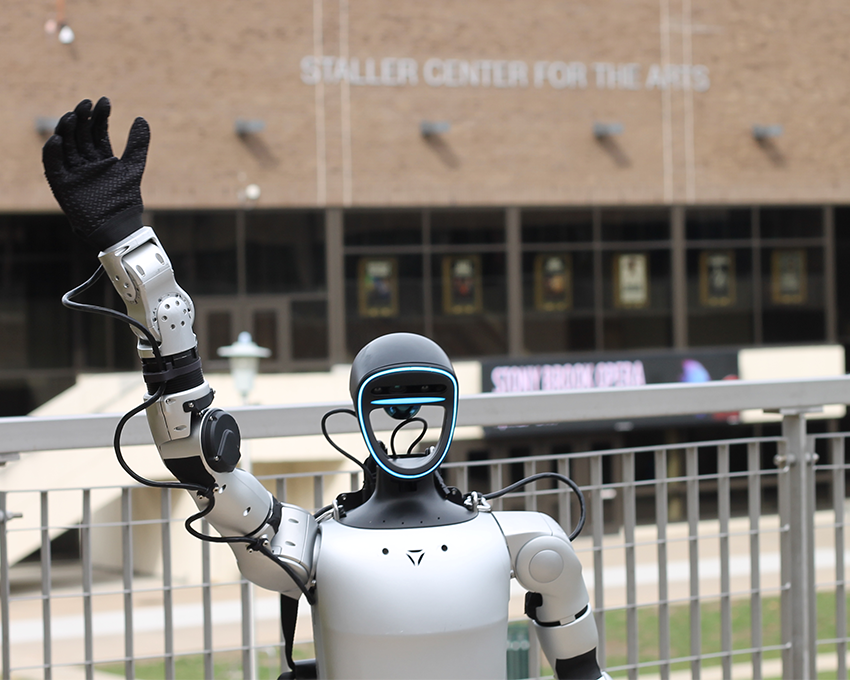 A humanoid robot with a sleek design waves its arm in front of the Staller Center for the Arts, which features a brick façade and visible posters in the background.