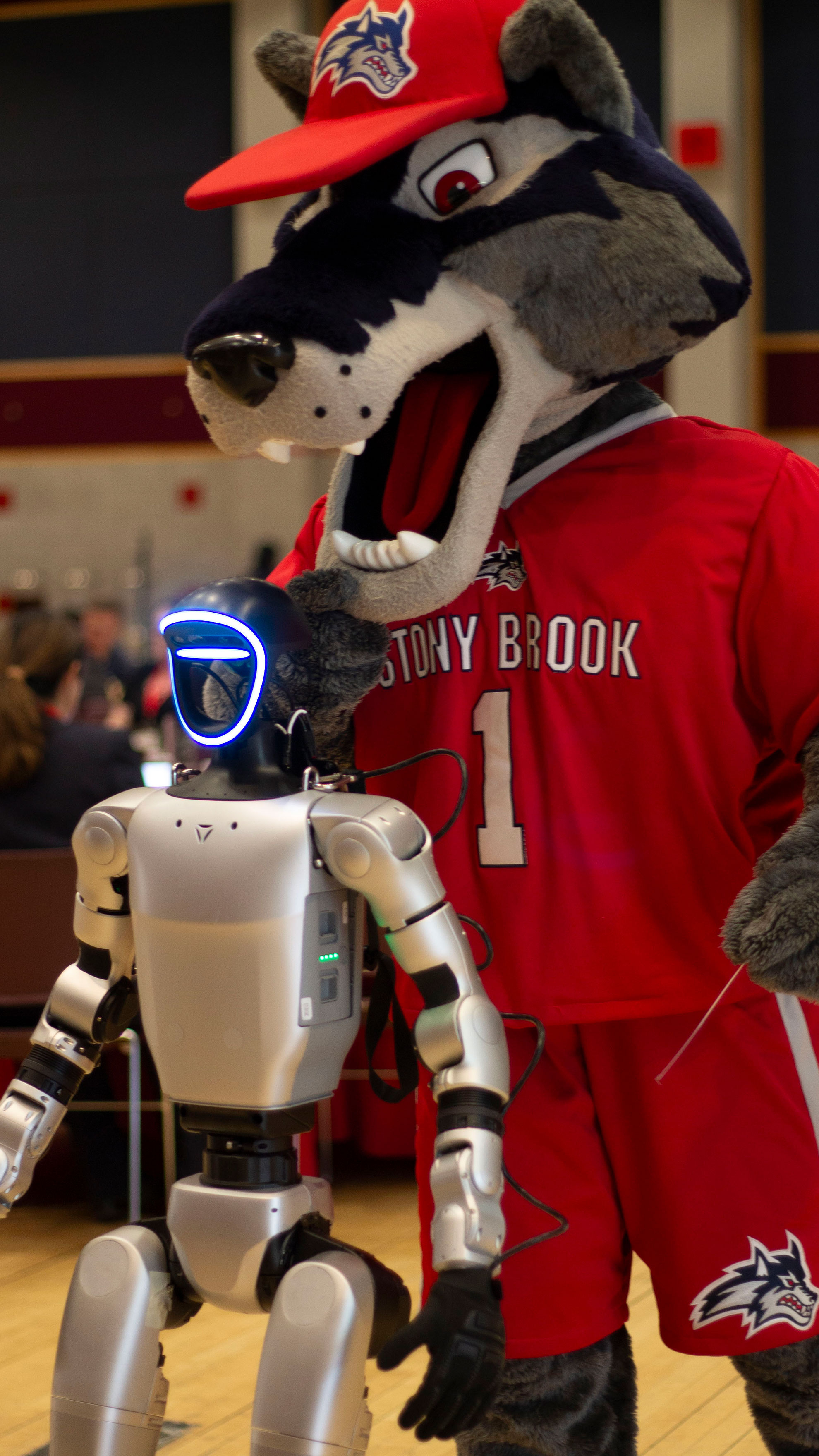 Wolfie the Seawolf mascot in a red jersey and cap stands next to MEC A Wolf, a silver humanoid robot with a blue-glowing face in a crowded setting.