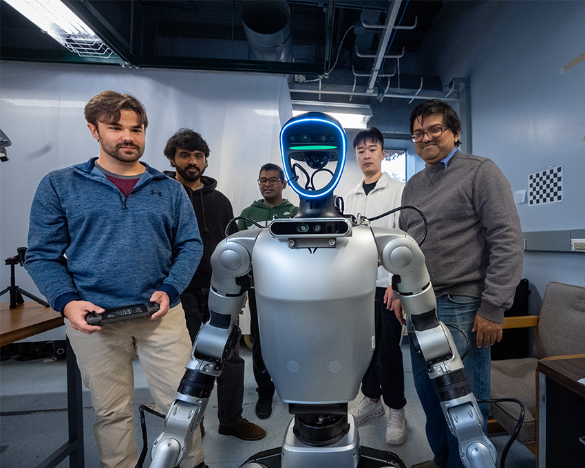 A group of five people, including a humanoid robot, in a modern workspace. The robot has a silver body and blue LED lights on its face, while the individuals are engaged around it.