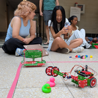 Several students sit on the floor during a hands-on robotics workshop, observing colorful, homemade robots on a taped course with toy obstacles. One person smiles while looking at a phone as others watch the activity.