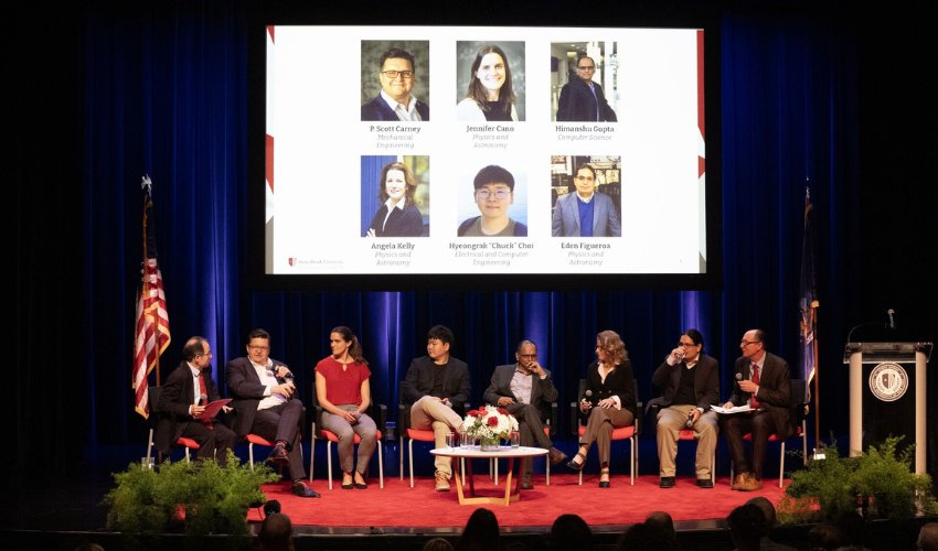 A panel discussion on stage with seven panelists seated in red chairs. A large screen displays headshots and names of the participants, alongside an American flag and a podium.