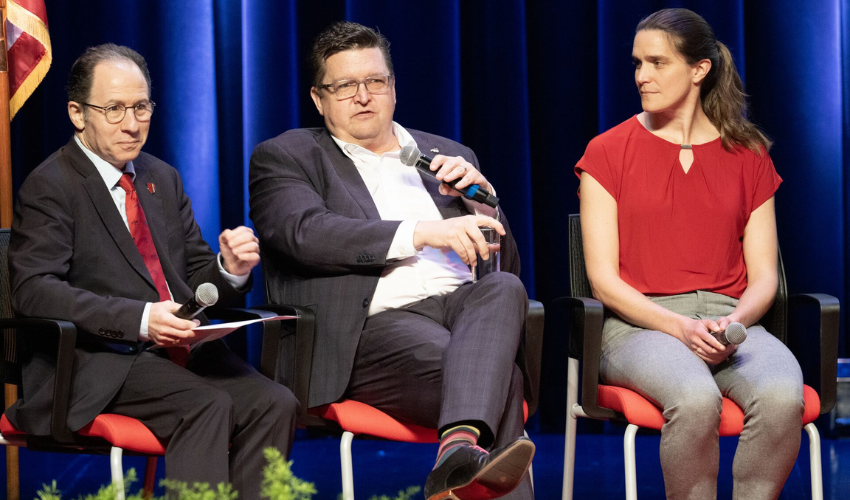 Three panelists are seated on stage during a discussion, with two men and one woman each holding microphones in front of a blue curtain