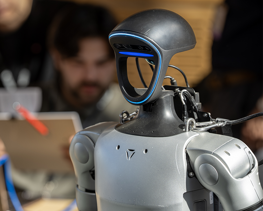 A humanoid robot with a sleek silver body and a black head with blue accents stands in the foreground, while blurred attendees are engaged in discussion in the background on a stairway.