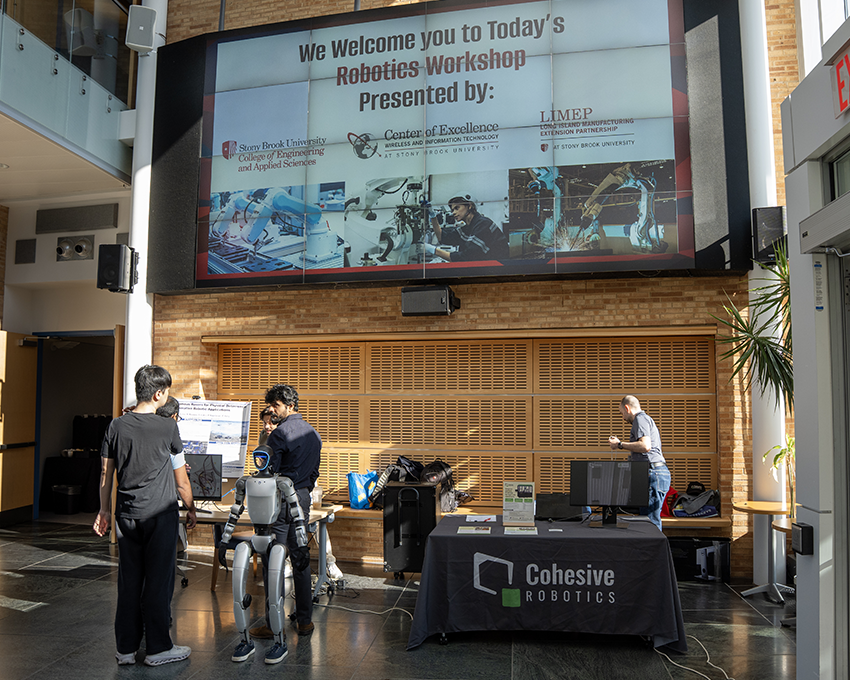 Attendees at a robotics workshop examine displays near a humanoid robot, with a large digital welcome sign in the background.