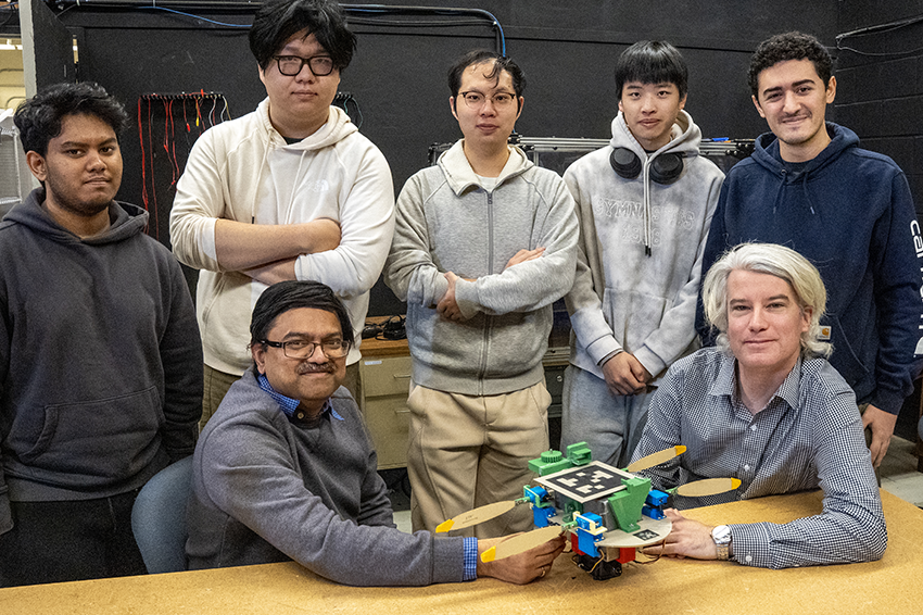 A group of seven people poses around a table in a lab with a crab-inspired air-to-water robot prototype. 
