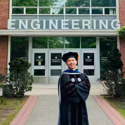 Yizong Li stands in a black cap and gown in front of the Engineering Building at SBU
