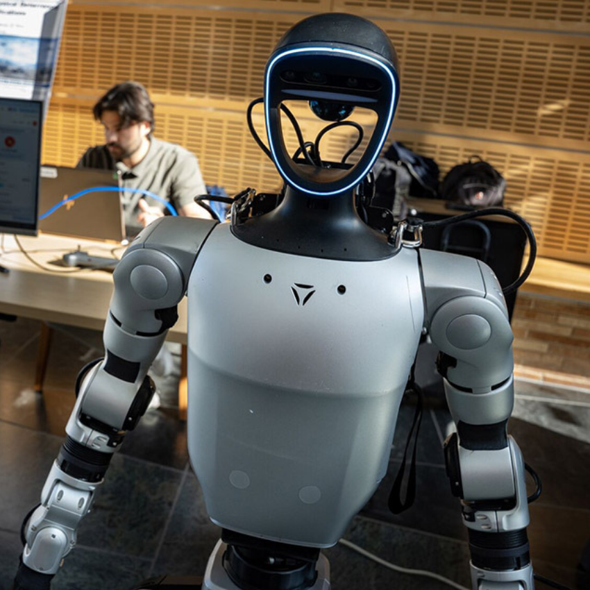 An image of a silver robot with a student working on a computer in the backgound
