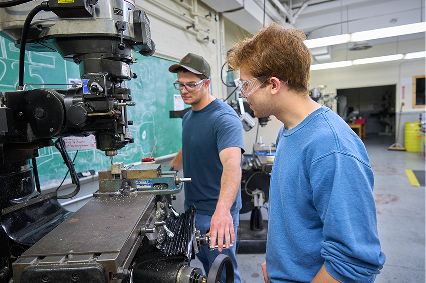 Two students in a workshop use a milling machine, one operating it and the other observing, both wearing safety glasses.