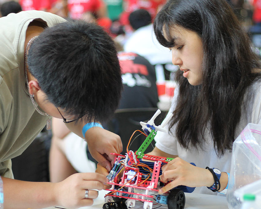 A young man and woman work together to assemble a small, colorful robotic device with wires and plastic parts while sitting at a table in a room.