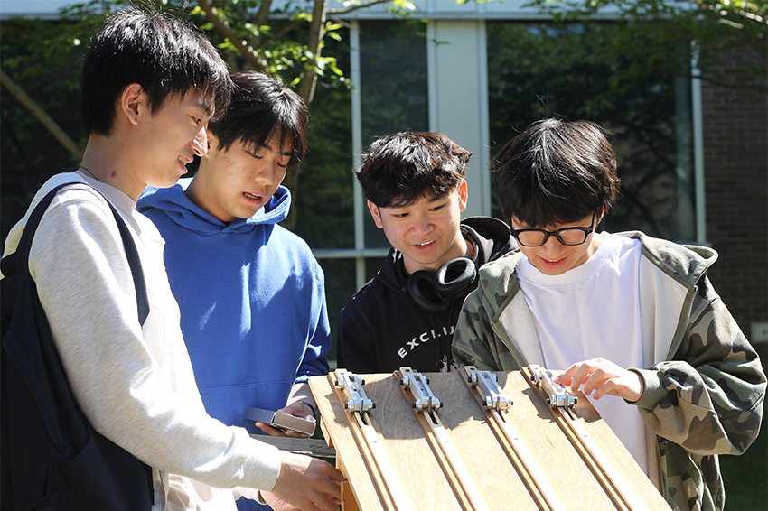 Four students gather around a wooden car racing track, enthusiastically examining their aluminum cars in an outdoor setting.