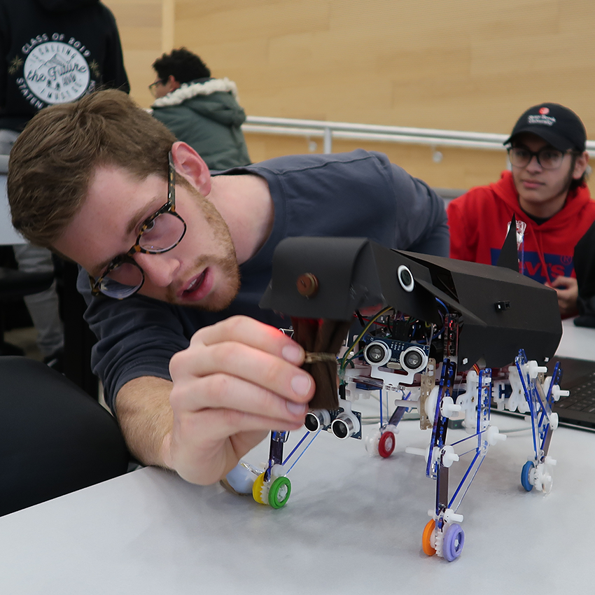 A young man with glasses adjusts a small, black, four-legged robotic creature on a white table while another student watches in the background.