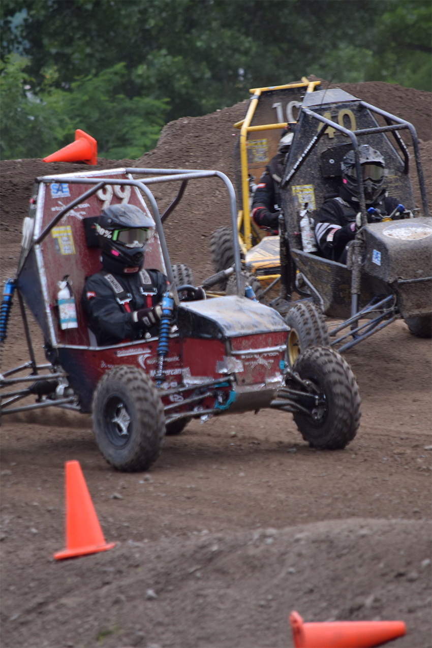 Two off-road racing vehicles maneuver through a dirt track, with one red vehicle in the foreground and one yellow vehicle behind it, both driven by masked racers.
