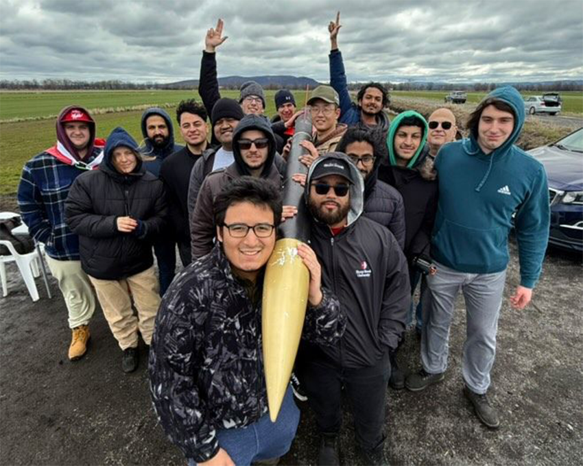 A group of students stands outdoors on a cloudy day, posing with a large, pointed rocket model held by two individuals in the center of the crowd.