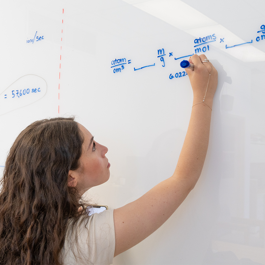 A young woman writes an equation on a white board with a blue marker