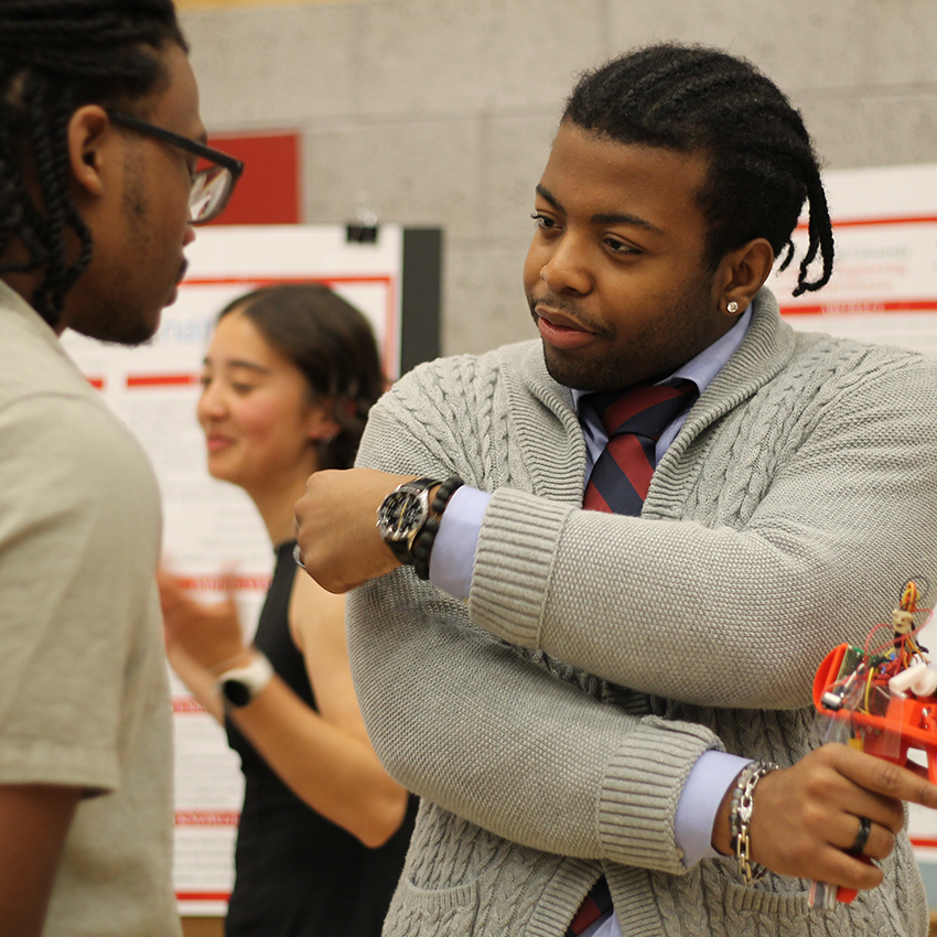 A student in a grey cable-knit sweater and tie talks to a peer while holding a small, orange medical device, with a woman visible in the background.