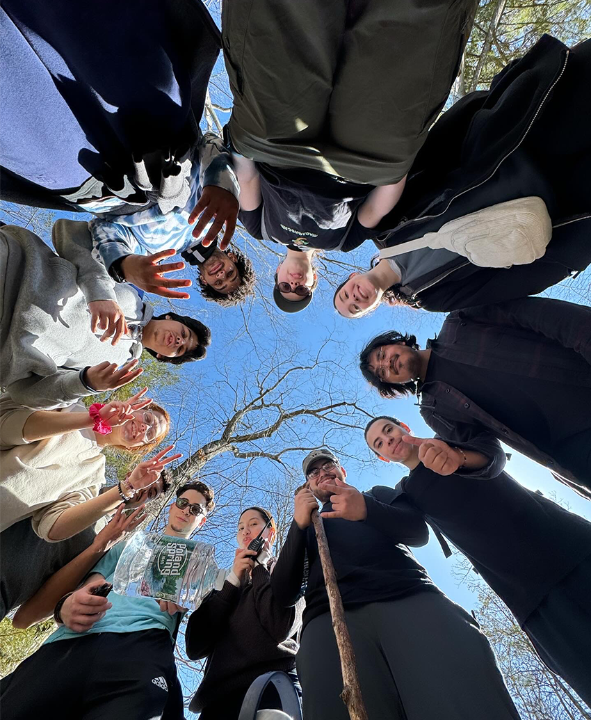 A diverse group of eleven individuals gathered in a circle, viewed from below, with a bright blue sky and tree branches above.