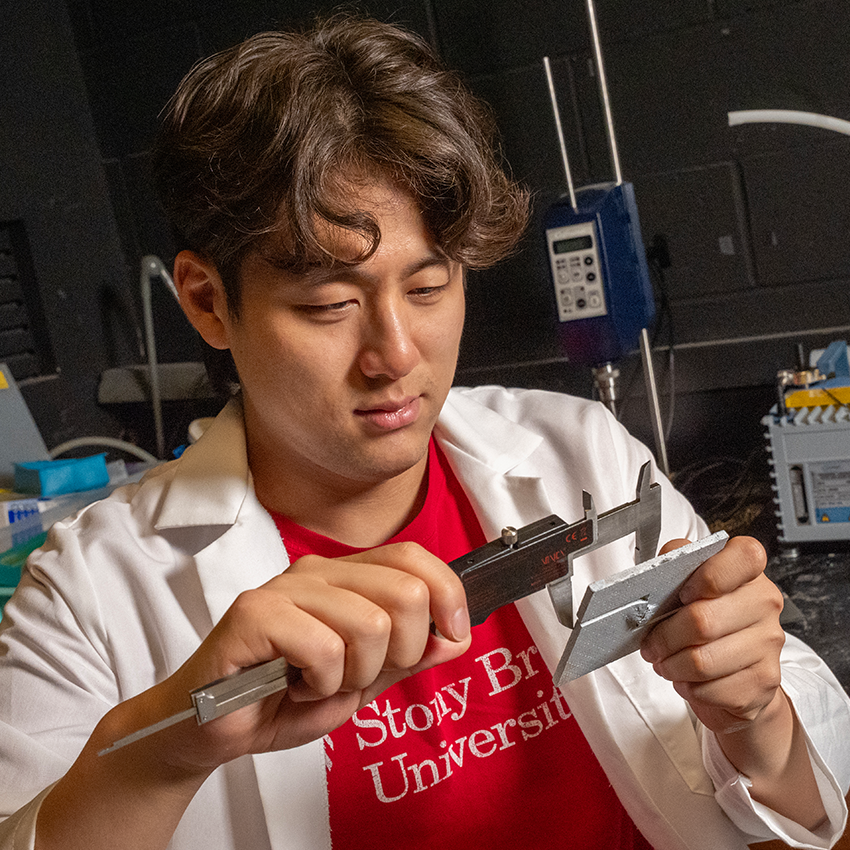 A student in a white lab coat and red Stony Brook University shirt uses a metal caliper to measure a small, square, grey textured material sample.