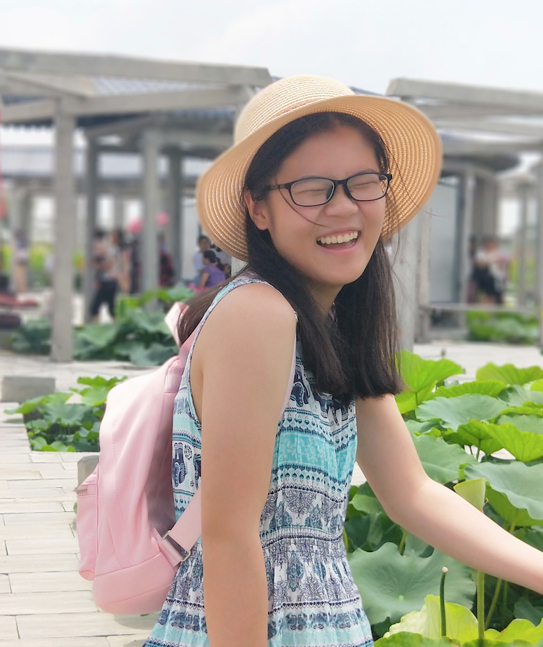 Portrait shot of Yilin Zhao smiling and wearing glasses, sunglasses, pink backpack and a blue patterned dress