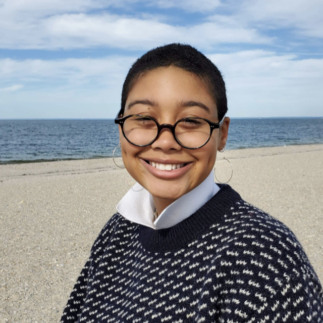 Close up shot of Braeden Bierwiler by the beach shore, wearing circle glasses and a navy sweater with a white collar 