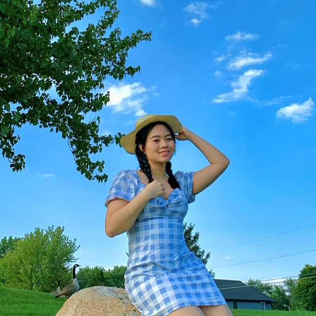 Medium shot of Stephanie Yam with pigtail braids, sitting on a rock wearing plaid blue dress and a beige hat