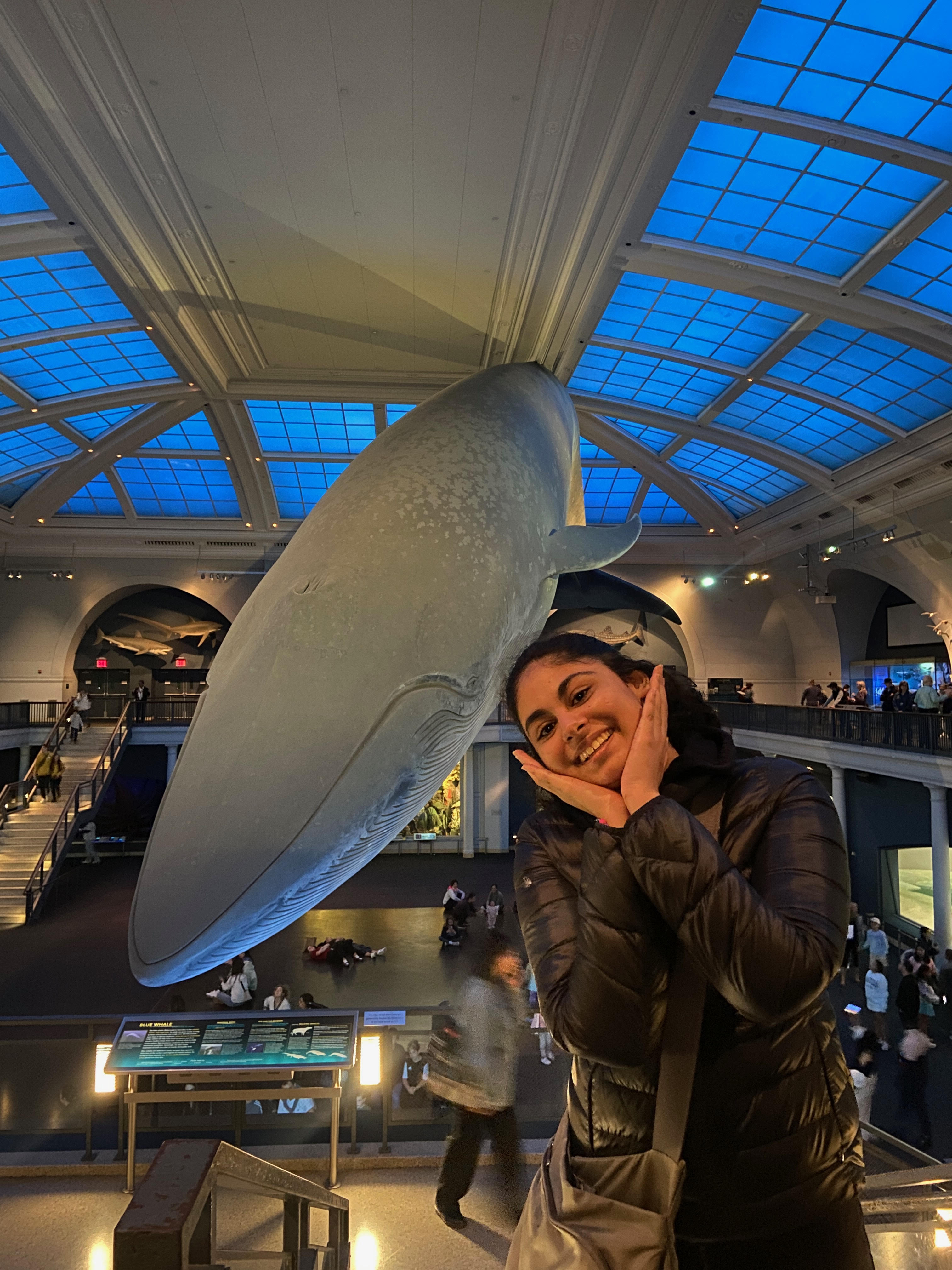 Medium shot of Maya with medium black wavy hair, wearing a black jacket psoing in front of a whale statue