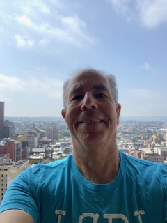 Micheal Newman smiling wearing a blue shirt and taking a selfie with a city and blue sky background