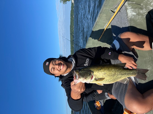 Camron wearing a black jacket and black cap, sitting on a boat while holding up a fish