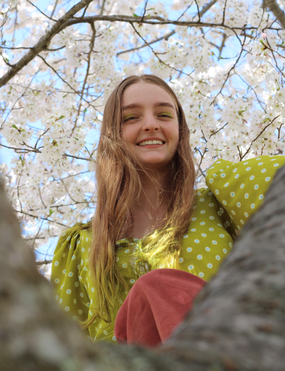 Medium close up shot of Isabella with blonde hair wearing a polka dot green shirt and smilling behind a tree
