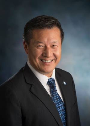 Headshot of Jun Liu Wearing a Black Suit with Blue Pattern Tie Against a Dark Background 