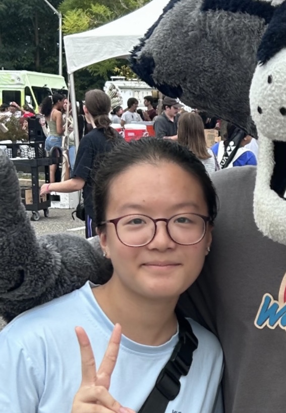 Wen Feng with circle glasses wearing a blue shirt and holding the peace sign while posing with the Stony Brook mascot wolfie