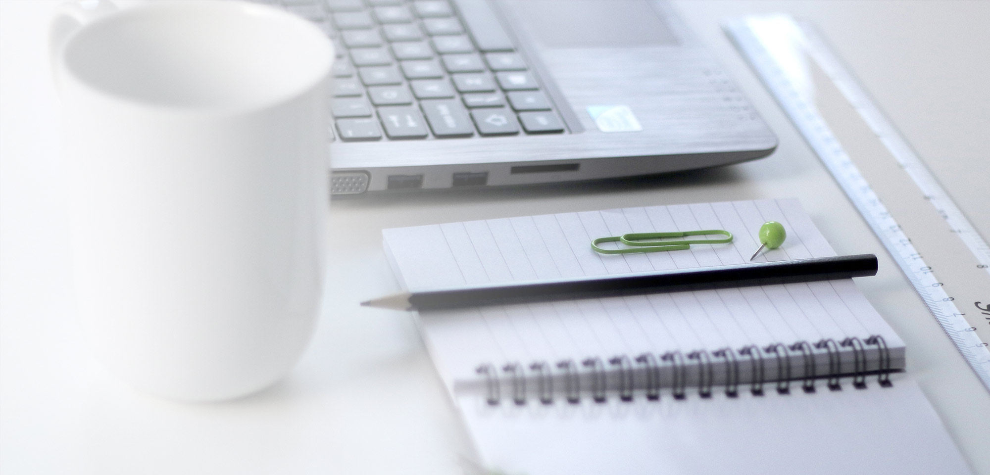 A laptop keyboard, spiral notebook, pencil, and coffee mug on a white desk
