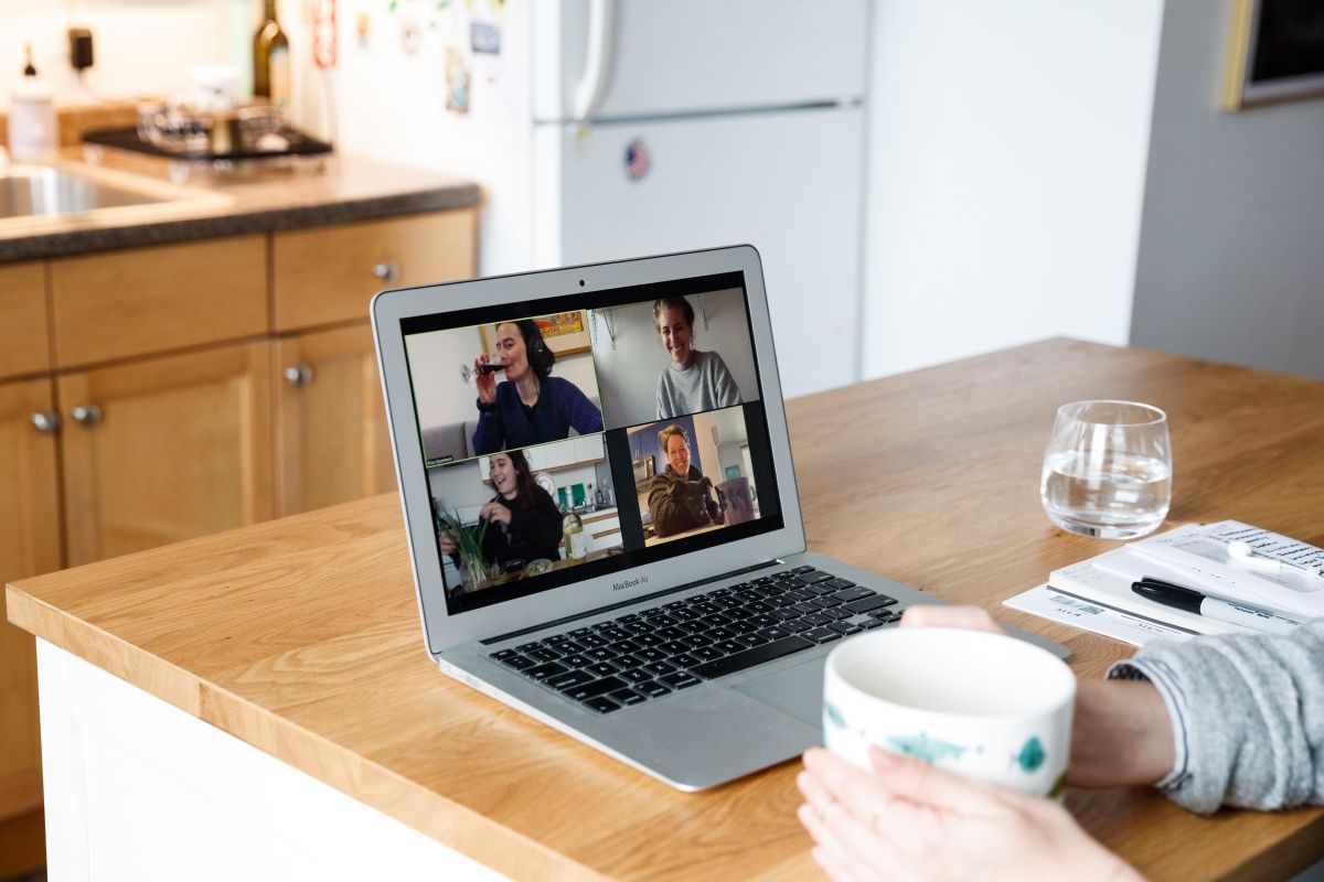 A person participating in a virtual meeting on a laptop, with four participants visible on screen in a kitchen setting